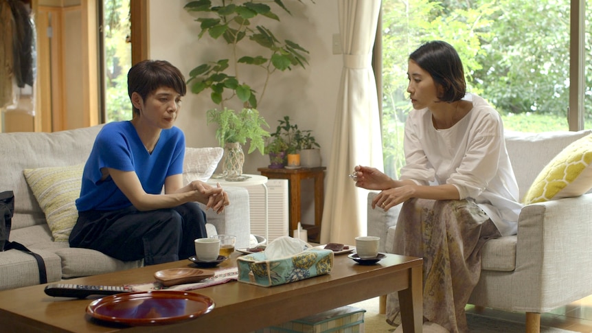 Japanese woman with cropped dark hair wears blue shirt and sits on couch next to Japanese woman with dark bob and white shirt.