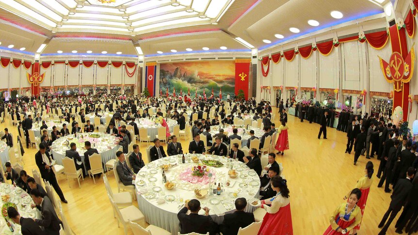 Men in suits sit at a large number of tables in a big banquet hall. They are being served by women in traditional Korean dress.