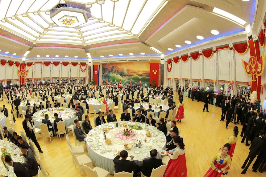 Men in suits sit at a large number of tables in a big banquet hall. They are being served by women in traditional Korean dress.