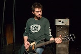 Steve Albini plays a guitar on stage, he wears a shirt with the word SNUB on it. 