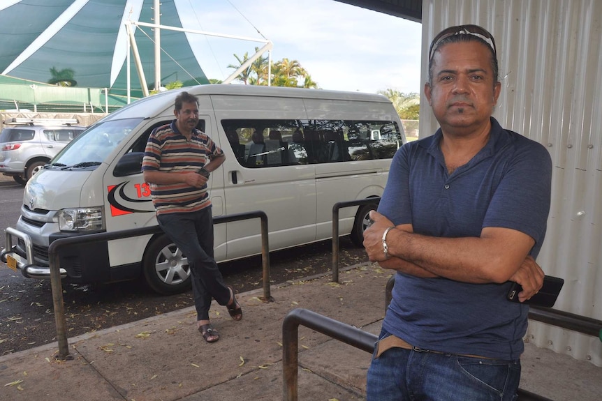 Two men stand in a taxi rank. One is closer to the camera with his arms folded across his chest.