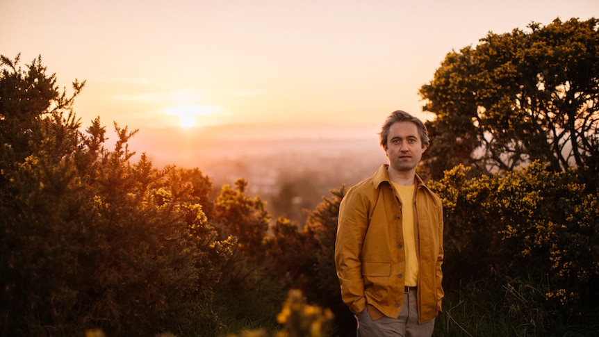 Conor O'Brien stands in front of a sunset and a thicket wearing a gold and yellow casual outfit
