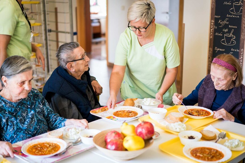 A staff member serves food to a group of nursing home residents at a dining table.