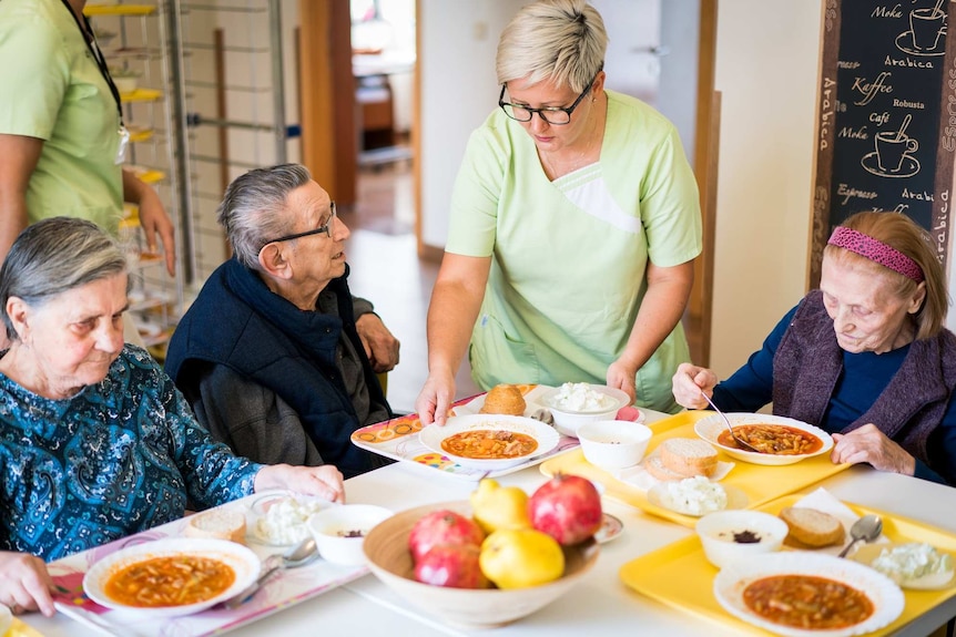 A staff member serves food to a group of nursing home residents at a dining table.