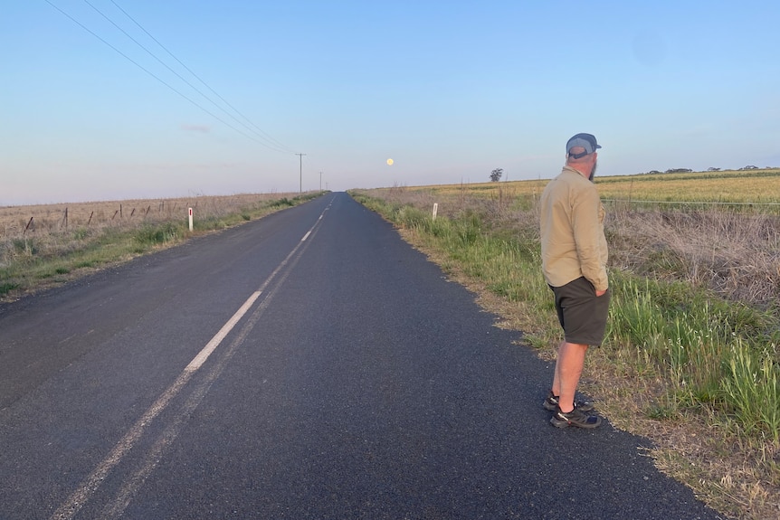 A man stands next to a long flat stretch of road in open countryside, with the moon in the early evening sky.
