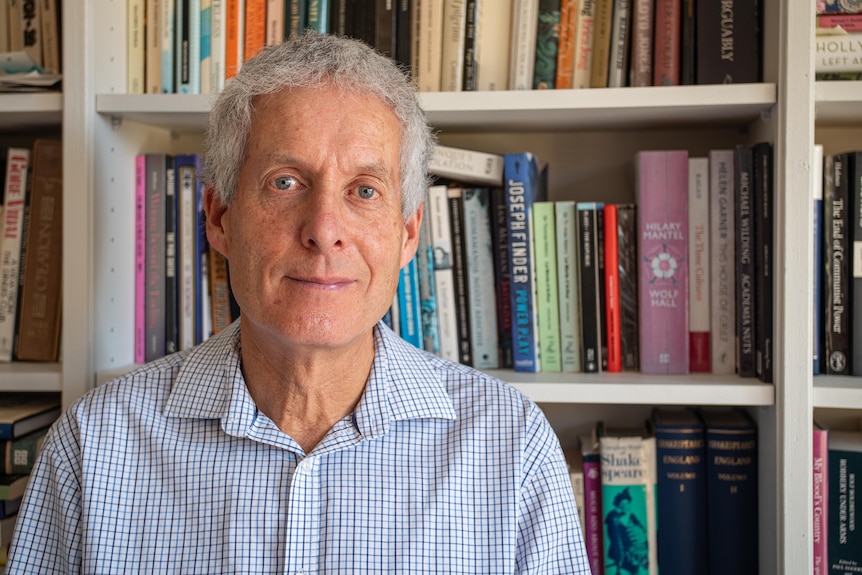 A man wearing a striped shirt sits in front of a bookshelf.