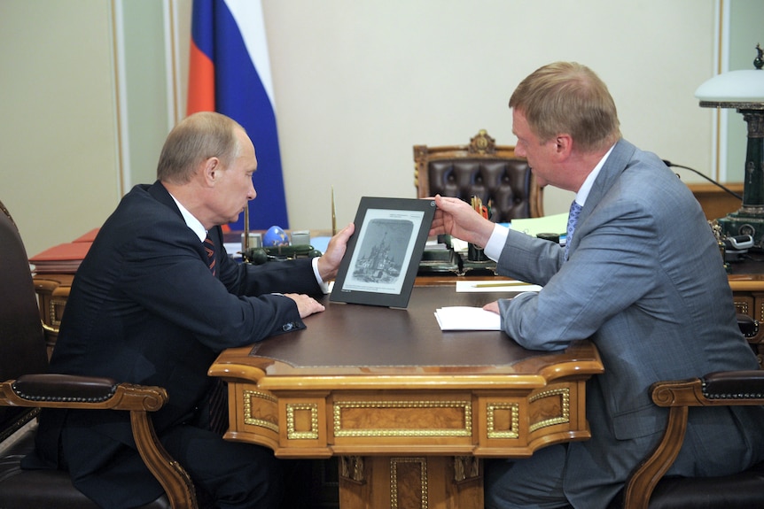 两个西装革履的男人面对面坐在一张装饰精美的办公桌前，手里拿着一个平板电脑原型。 俄罗斯国旗在左侧背景中可见
