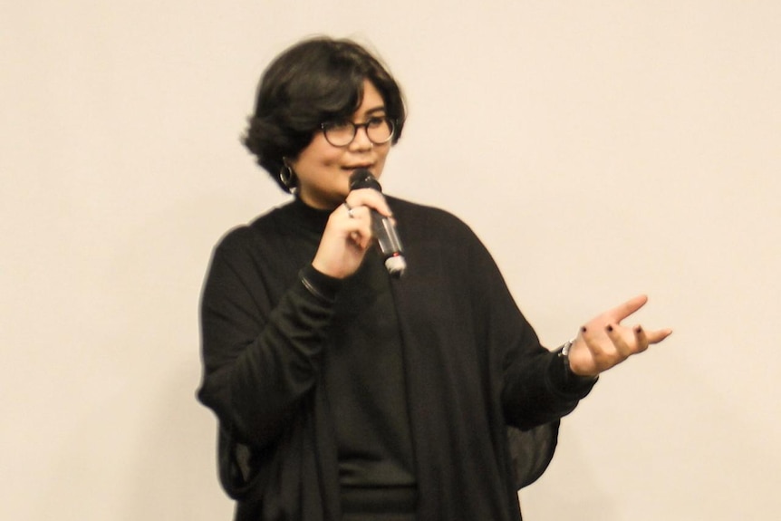 A woman in a black outfit stands in front of a cream wall and speaks into a microphone.