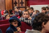 A child leans over a chair as he sits among a crowd of other children.