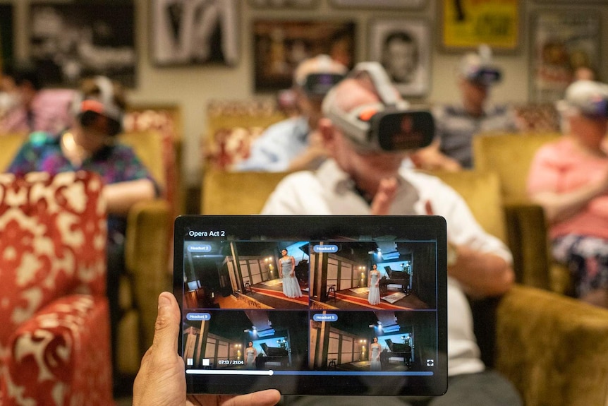 A tablet in the foreground shows an opera singer and in the background six older people wear VR goggles.