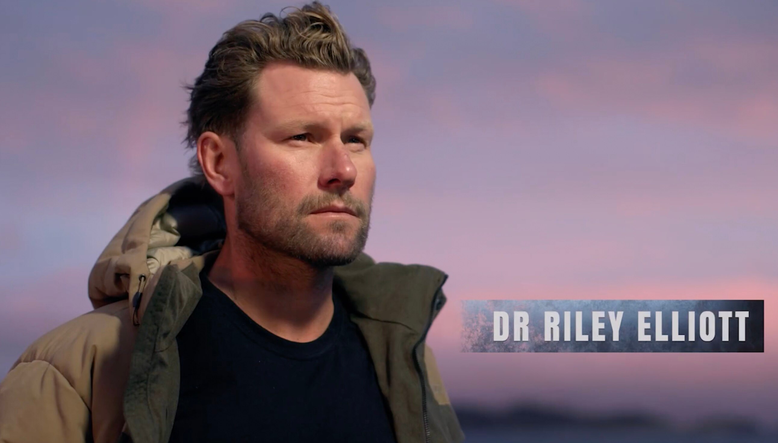 A man looks off into the distance, wearing a green jacket and black shirt.