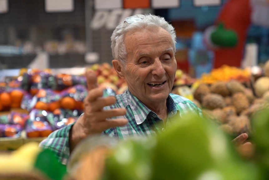 An older man with grey hair and glasses on his head, smiles while looking at fruit and vegetables.