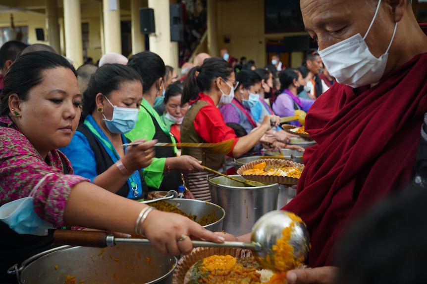 A row of people scoop servings of food out of large pots. Many are wearing surgical masks