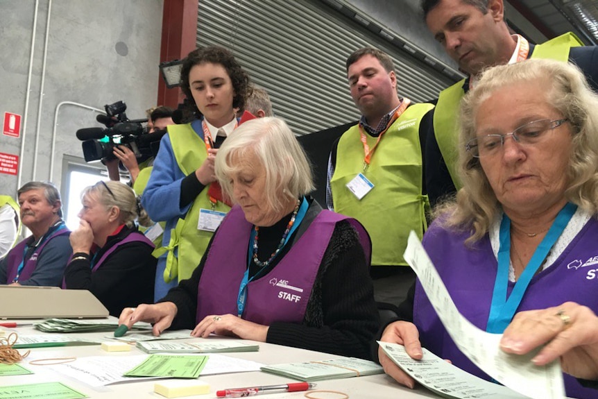 Australian Electoral Commission staff in purple vests sit at a table counting votes as monitors in green vests watch over.