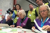 Australian Electoral Commission staff in purple vests sit at a table counting votes as monitors in green vests watch over.
