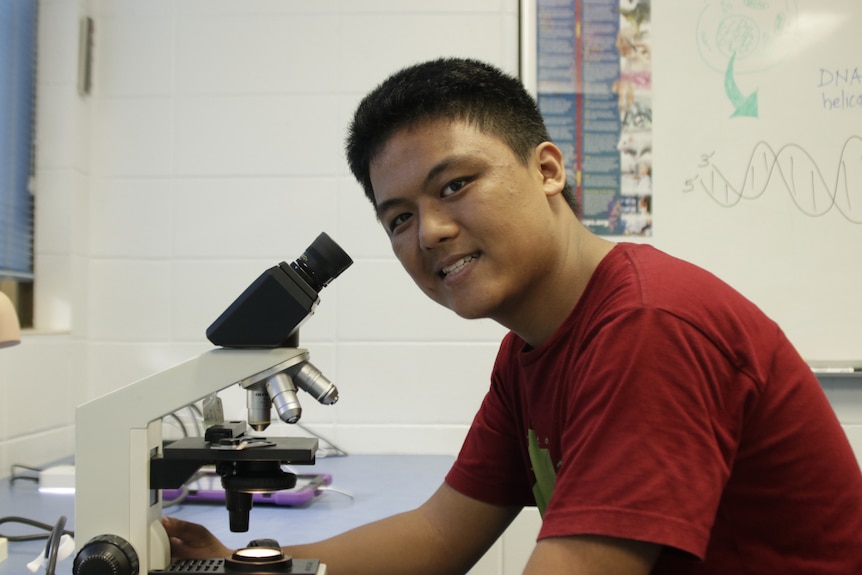 A teenage boy sitting at a table, next to a microscope, while looking at the camera and smiling.