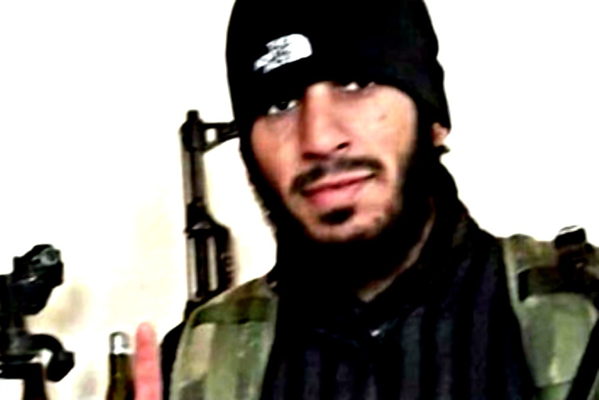 A bearded man in a beanie and camouflage vest standing in front of a rifle