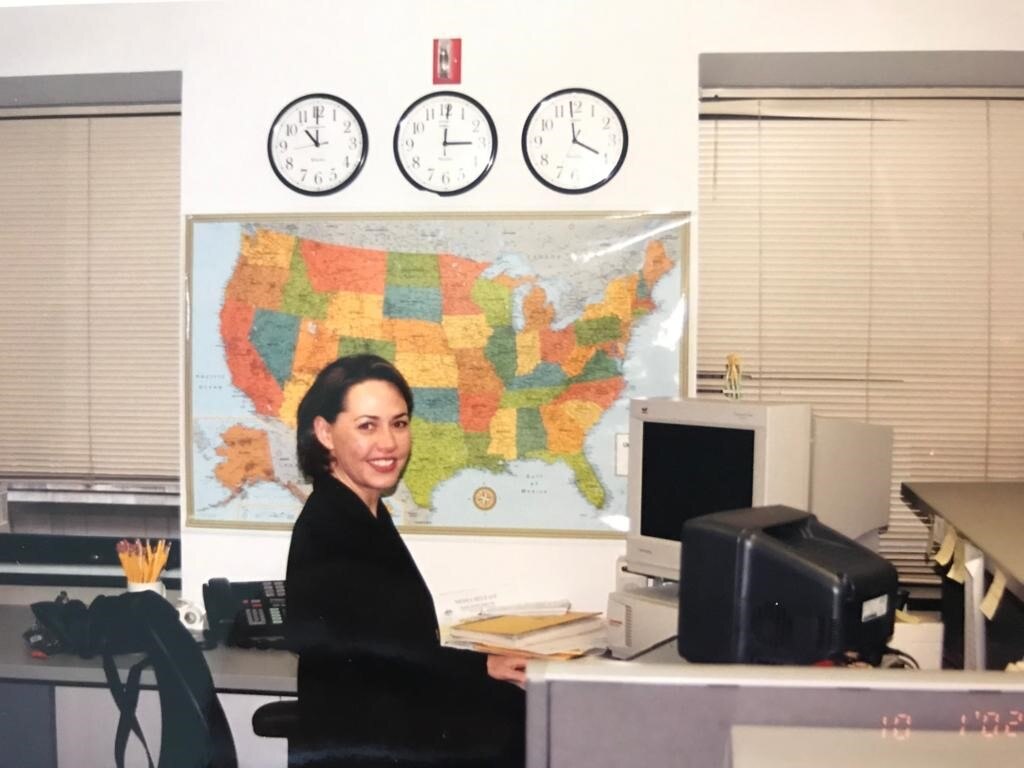 Woman sitting at desk with old fashioned computer and map of USA and clocks on wall.