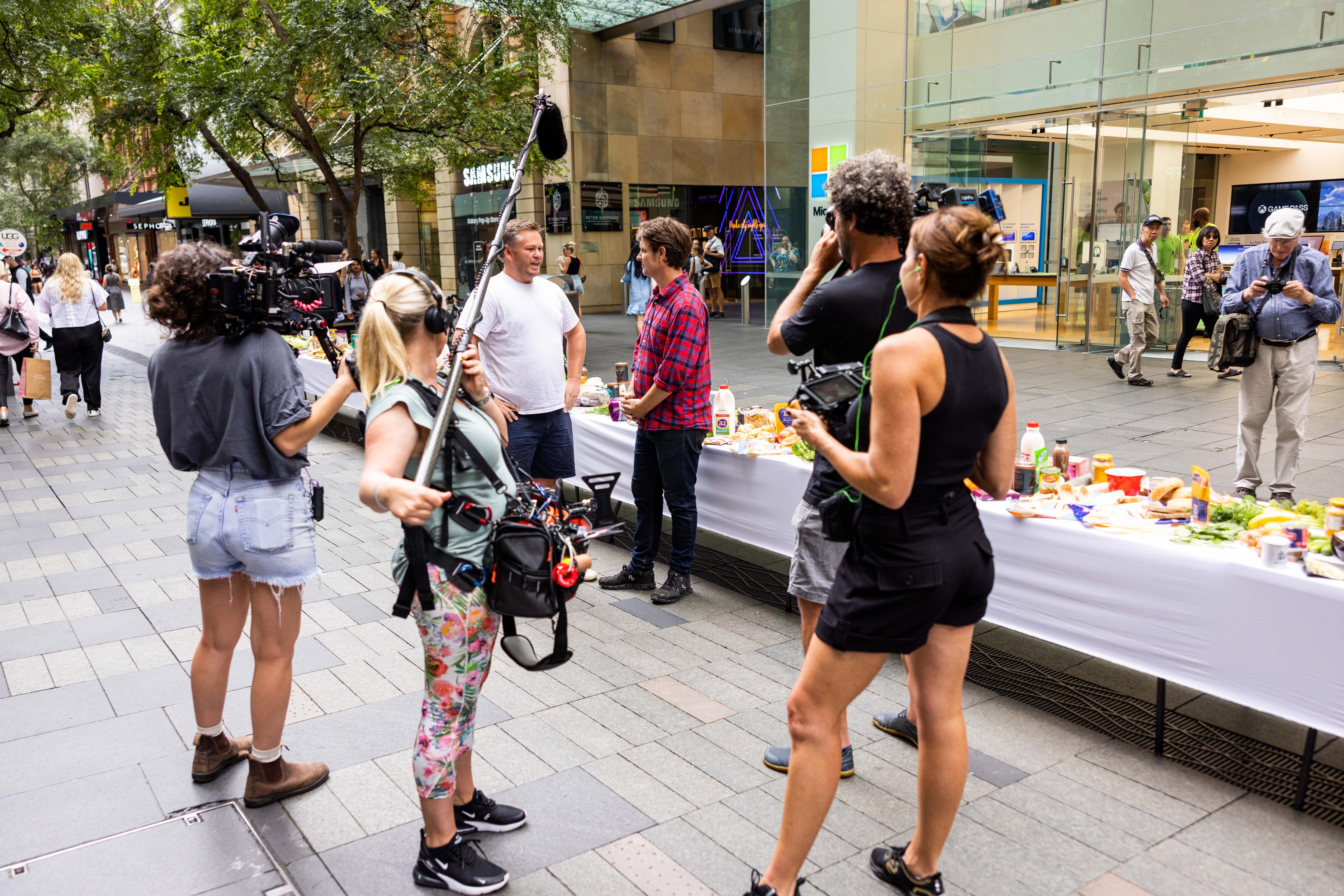 Camera crew filming two men talking while standing next to long table with food on it in mall.