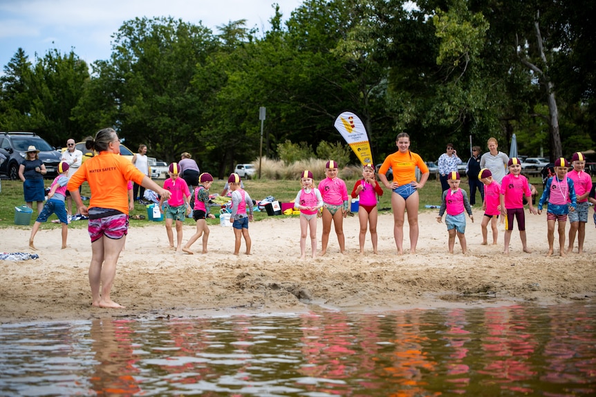 Several kids in swimming caps and pink shirts line up on the beach of Lake Canobolas alongside two adults in orange shirts.