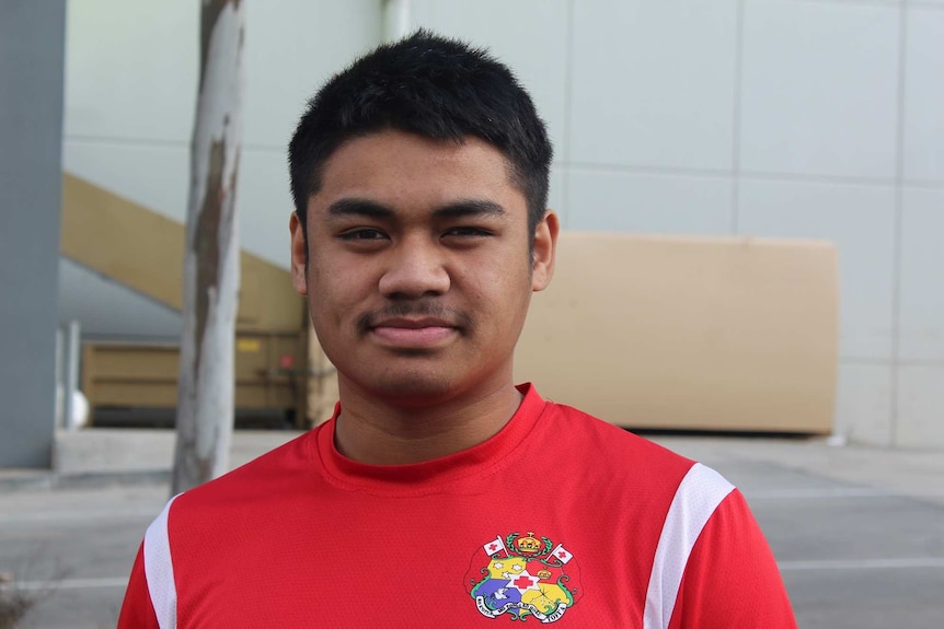 A boy with cropped black hair and wearing a red t-shirt has a serious expression on his face.