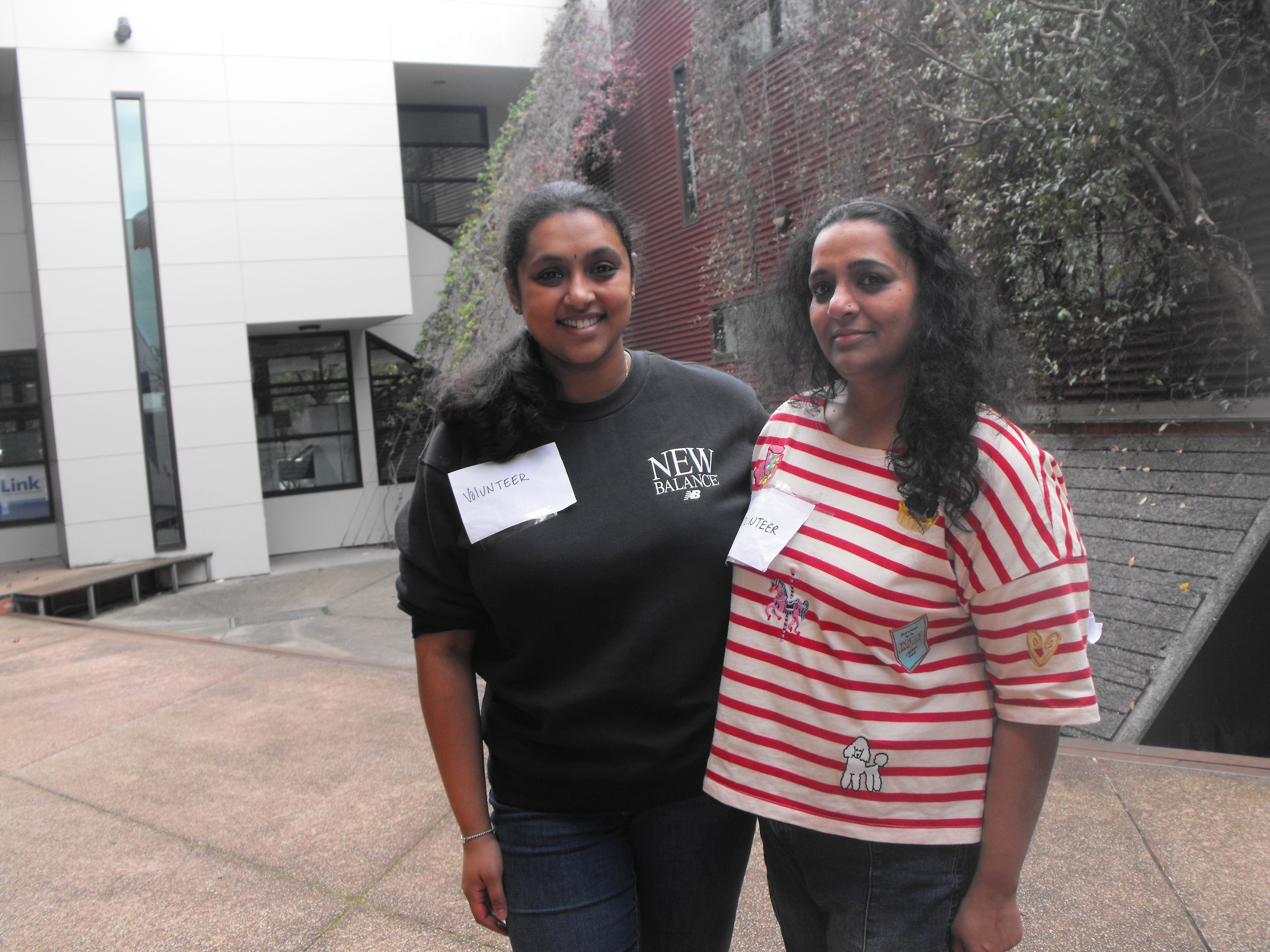 Gayathri and Varshini stand close together in a courtyard, both smile ,white paper with volunteer pinned to chest.