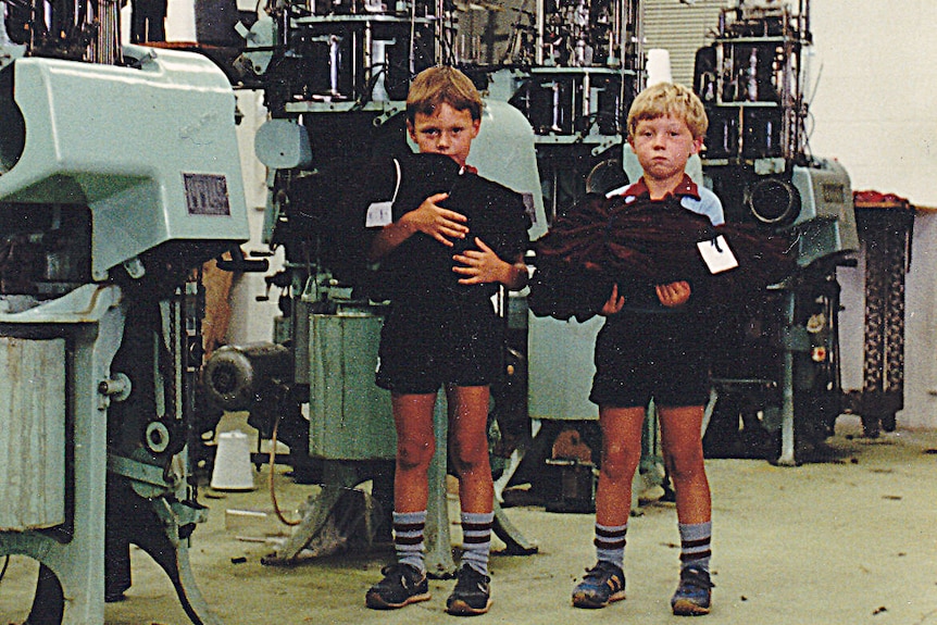 Two young boys hold piles of material next to machinery in a factory