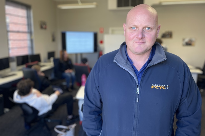 A bald man stands in a classroom, looking at the camera.