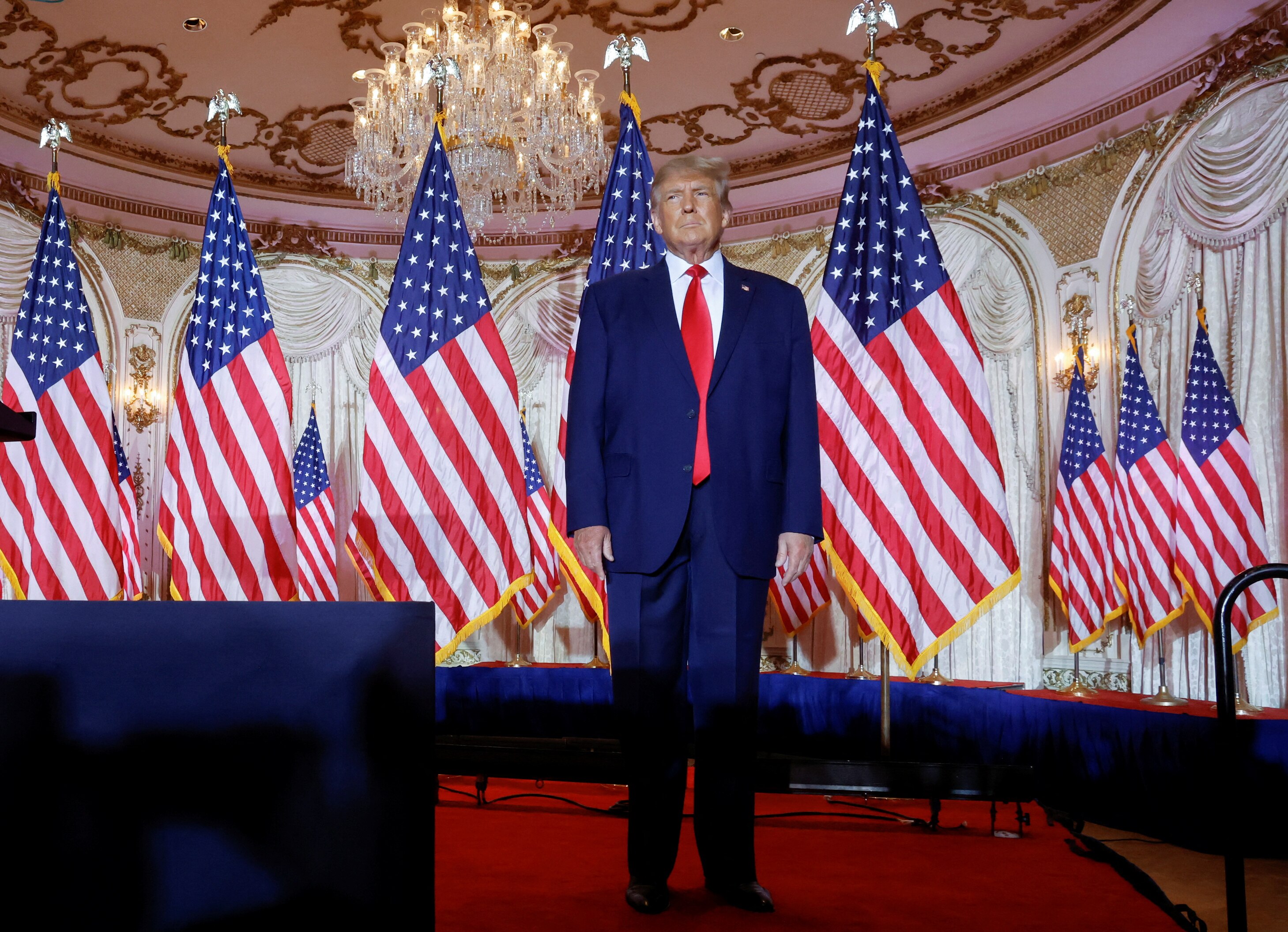 Donald Trump stands on an empty with is hands by his sides in front of several American flags