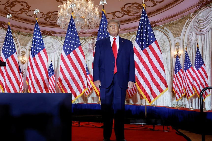 Donald Trump stands on an empty with is hands by his sides in front of several American flags
