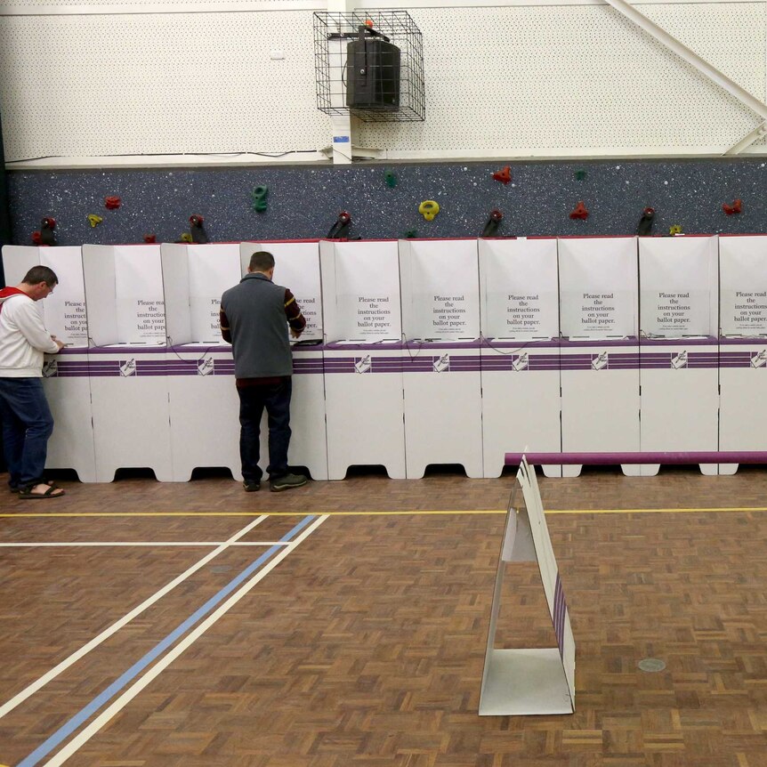 Three people fill in their voting forms at a long line of voting boxes.