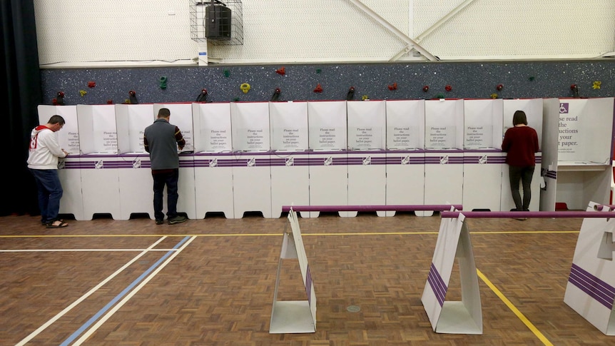 Three people fill in their voting forms at a long line of voting boxes.