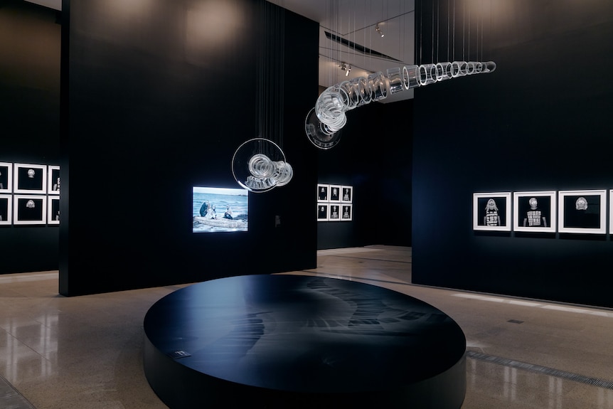 A black gallery space, with a glass eel trap sculpture hanging from roof, framed photographs and video projection on wall