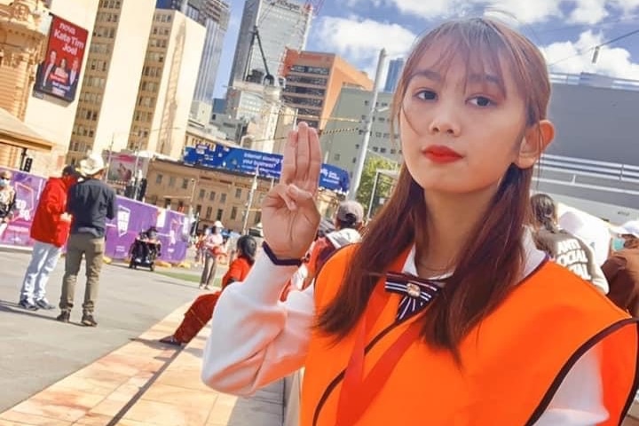 A young woman in a work uniform and high-vis vest raises three fingers on her right hand as she sits in a sunny public square.