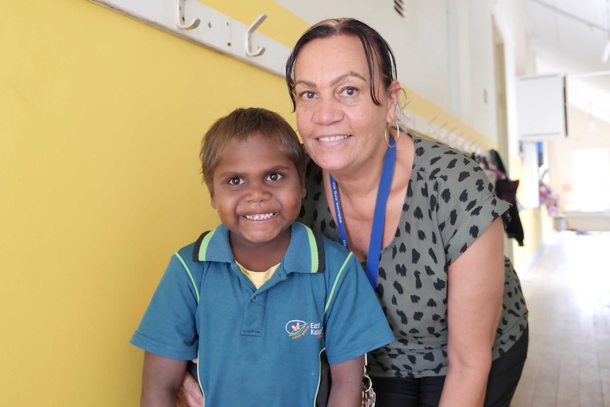 Indigenous student in uniform grins as woman leans forward smiling next to him. Both stand in yellow school hallway.