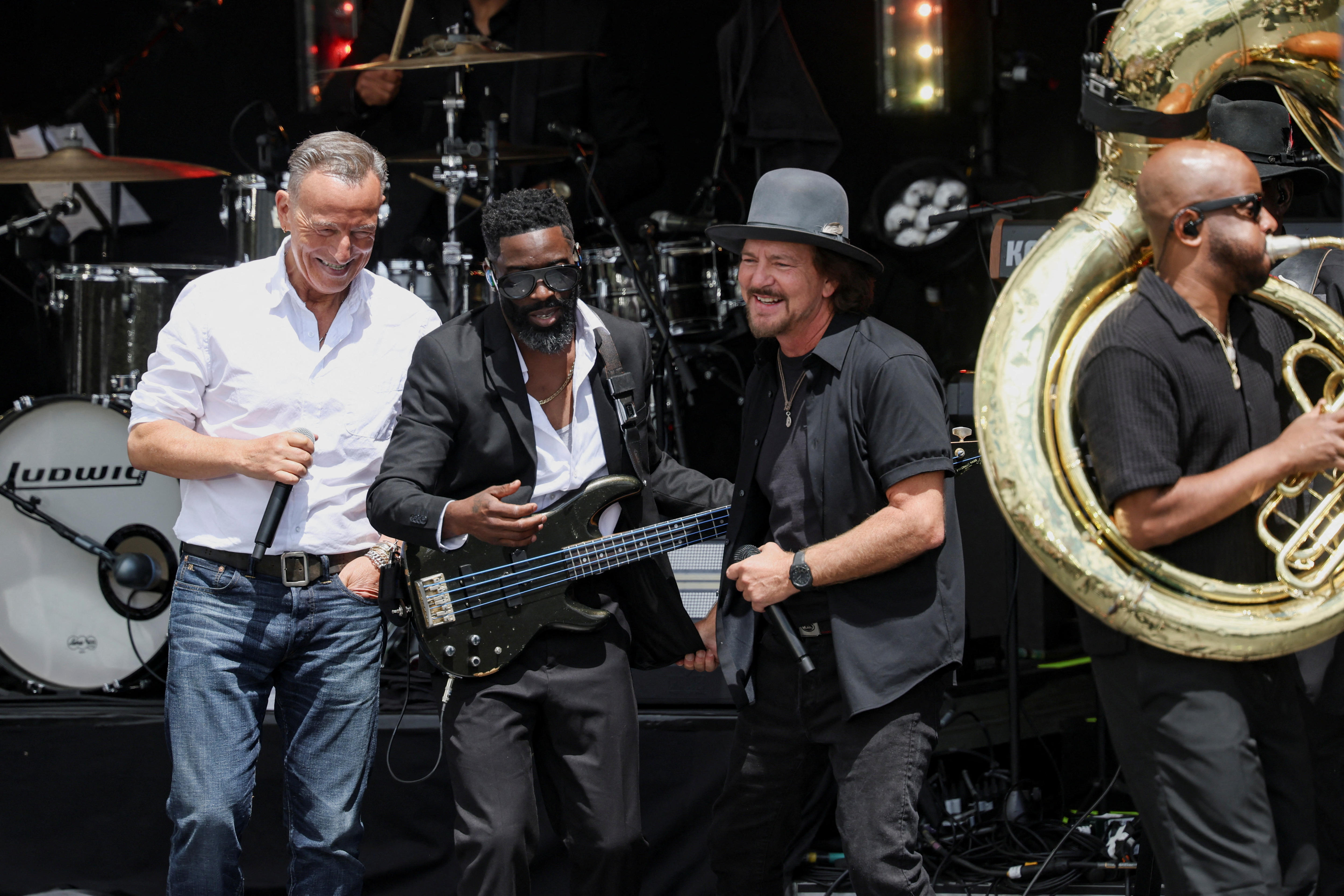 Four male musicians and singers on stage, one with a guitar and one with a sousaphone