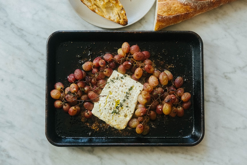 A block of feta cheese and red grapes, roasted on a tray with thyme to create a simple meal when served with a baguette