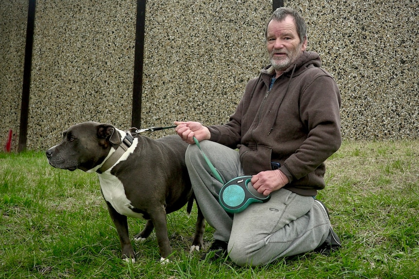 Graham wears a brown hoodie and grey tracksuit pants. He kneels next to a brown and white dog, which is on a leash.