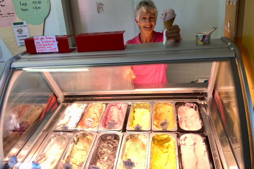 A lady holds up a cone of ice cream with colourful trays of ice cream in a fridge.