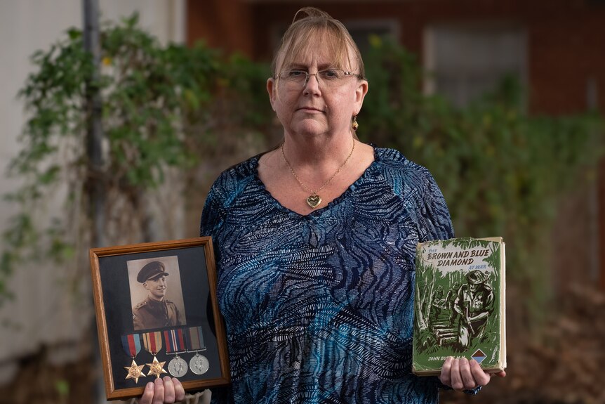 A woman holds up an old photograph of a soldier with medals underneath and a book in the other hand