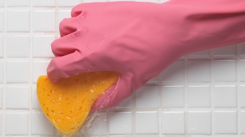 A close-up image of a hand with a rubber glove on it, holding a sponge which is being used to scrub tiles