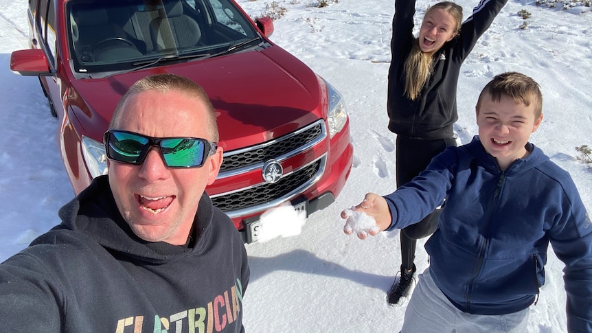 A man smiles at the camera in front of a red car. On the right a boy throws snow and a girl has her arms in the air.