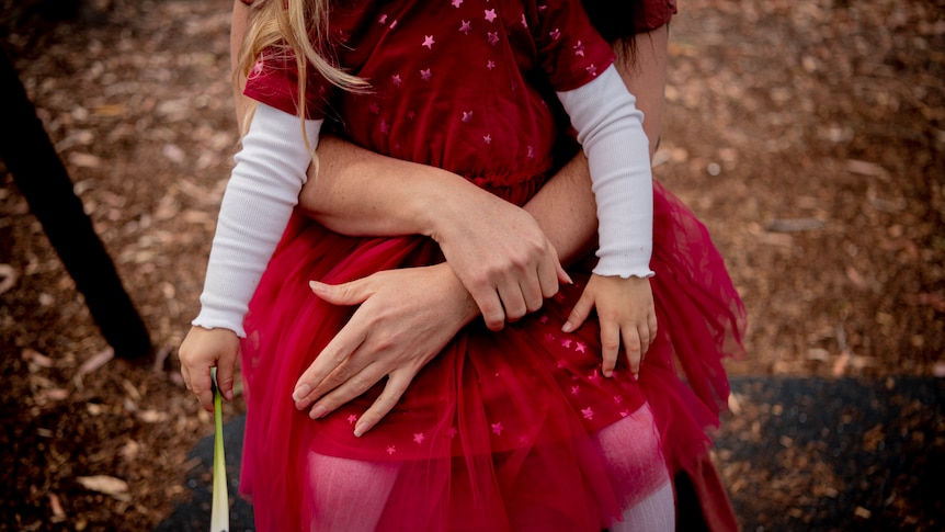 A mother holds her two-year-old daughter who is wearing bright red dress around the waist