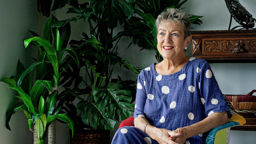 A woman in a navy blue dress with large white spots sitting in a chair inside a room and smiling.