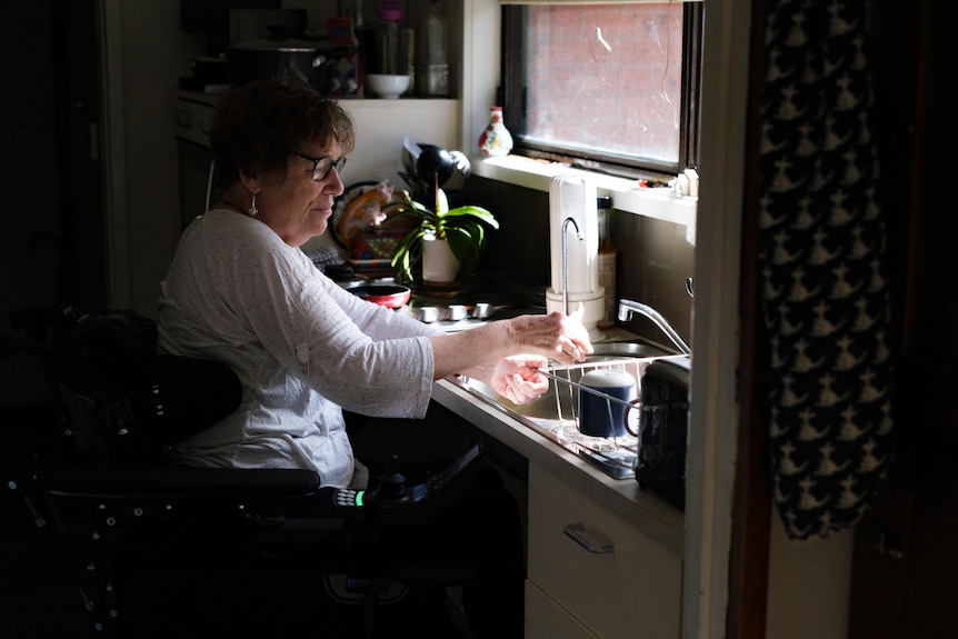 Deb washes the dishes at a sink with a window infront of her lighting her face.