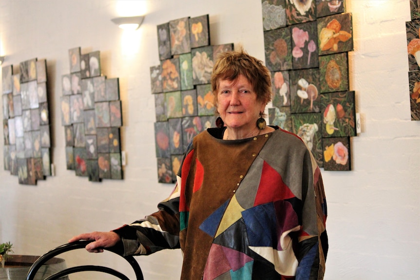 A red-haired woman, wearing jumper with large print, stands in front of a wall of paintings of colourful fungi.
