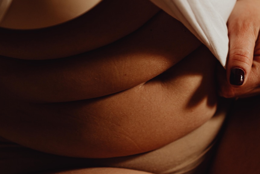 A close up photo of a woman with dark nail polish pulling a white top over her stomach
