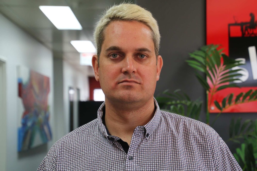 A head and shoulders shot of a man with short blonde hair standing in an office.