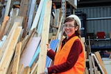 A women wearing a high-vis vest holds a piece of timber and smiles at the camera