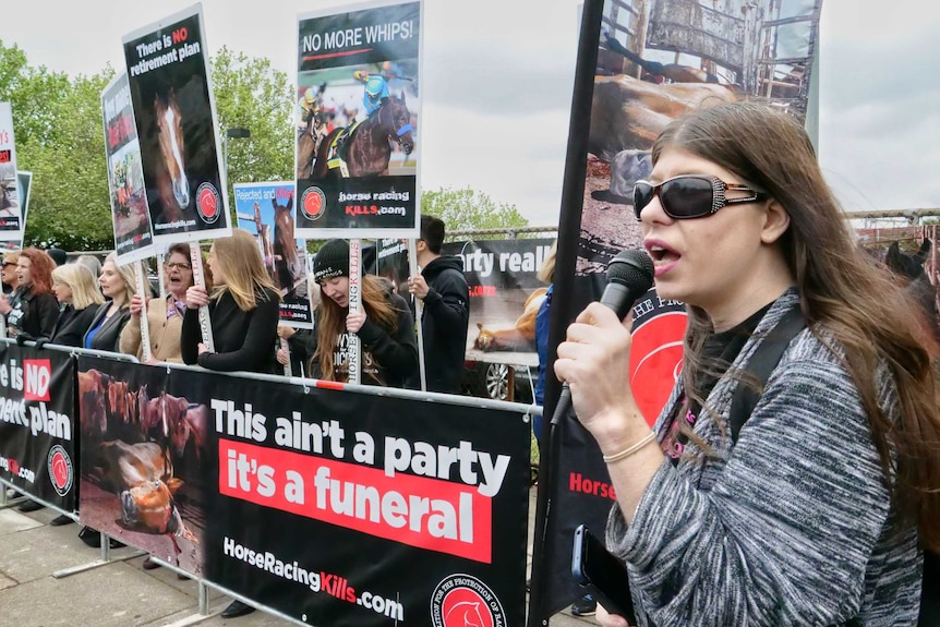 A woman wearing sunglasses and holding a microphone speaks while people hold horse-racing protest signs behind her.